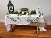 Decorated table with a white tablecloth, greenery, flowers, and a sign reading 'Into the Woods' on a wooden floor.